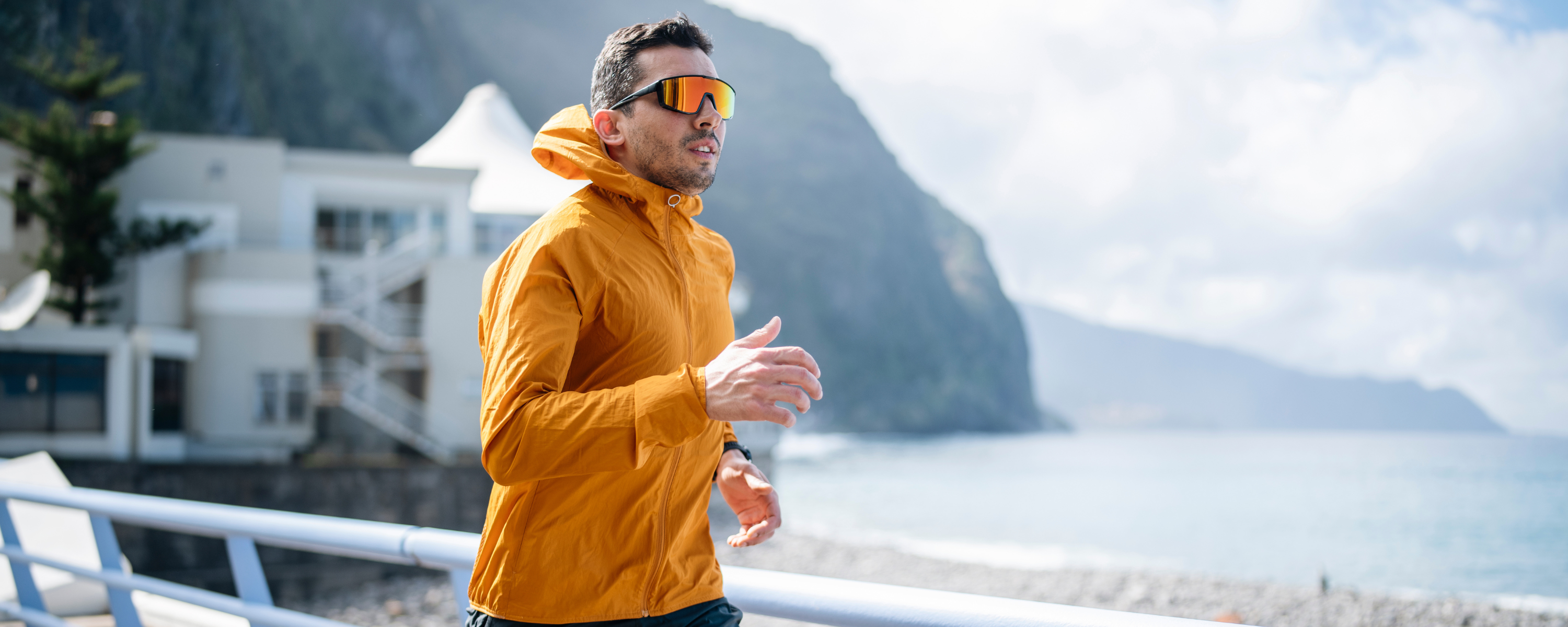 A man in a yellow jacket and Hokky running sunglasses standing on a coastal path with mountains in the background